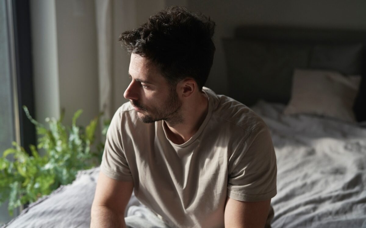 Man sitting on bed looking out window showing signs of testosterone and mental health struggles