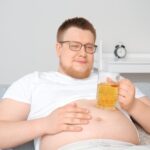 Overweight man sitting on couch holding a beer mug with visible belly fat representing how alcohol and weight loss work against each other