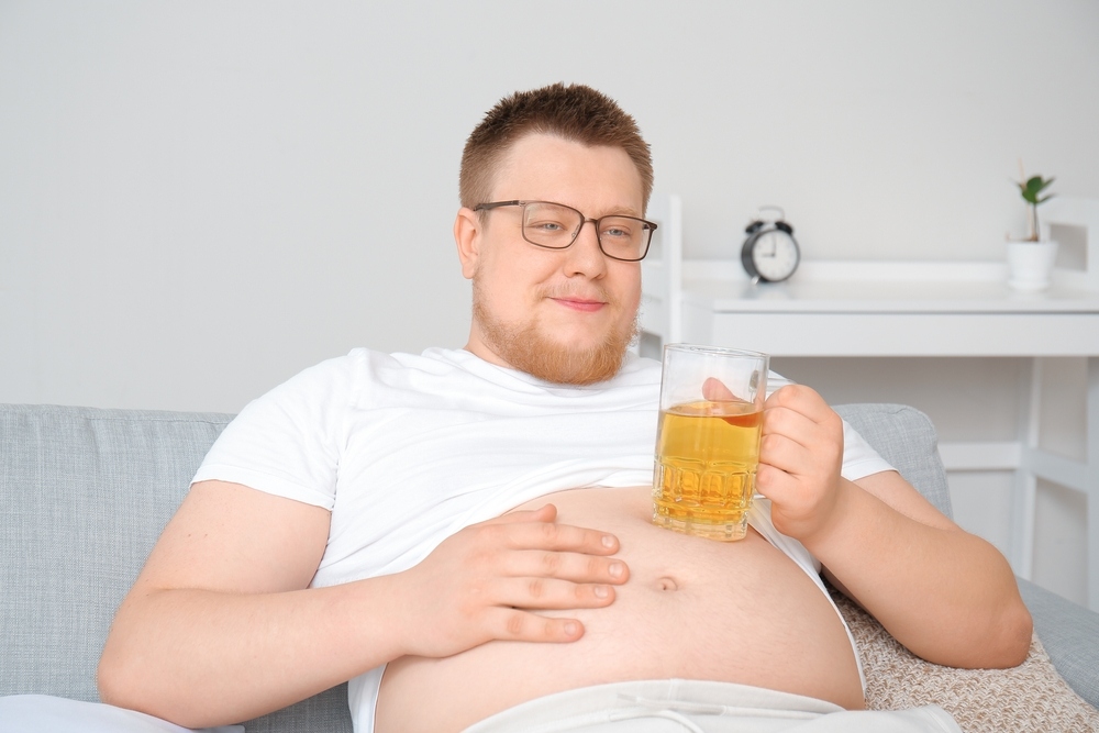 Overweight man sitting on couch holding a beer mug with visible belly fat representing how alcohol and weight loss work against each other